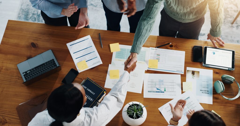 Overhead view of a diverse leadership team shaking hands over a conference table filled with financial charts and strategic planning documents.